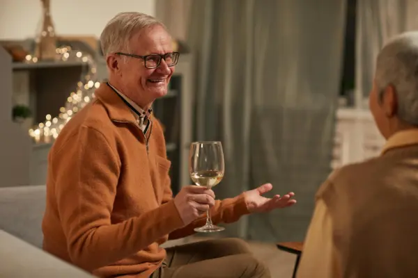 An older man smiling and talking while holding a wine glass on a couch, representing social recovery and post-prostatectomy pelvic floor rehabilitation at Monty Pelvic Physio in Montmorency.