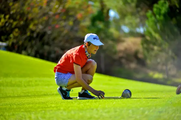 A woman wearing a red shirt and blue visor crouching on a green golf course to line up a putt, illustrating a return to community sport and lifestyle activities through prolapse management in Warrandyte.